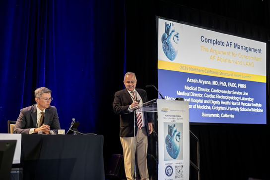 Speakers participate in a conference session on stage, with presentation slides displayed on a large screen in the background.